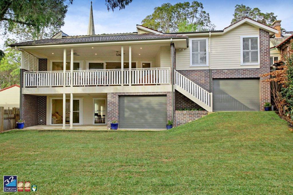 Two-story suburban house with a wraparound porch, brick and light-colored siding, double garage doors, exterior stairs, and a sloped grassy front yard. Trees surround the house in the background.