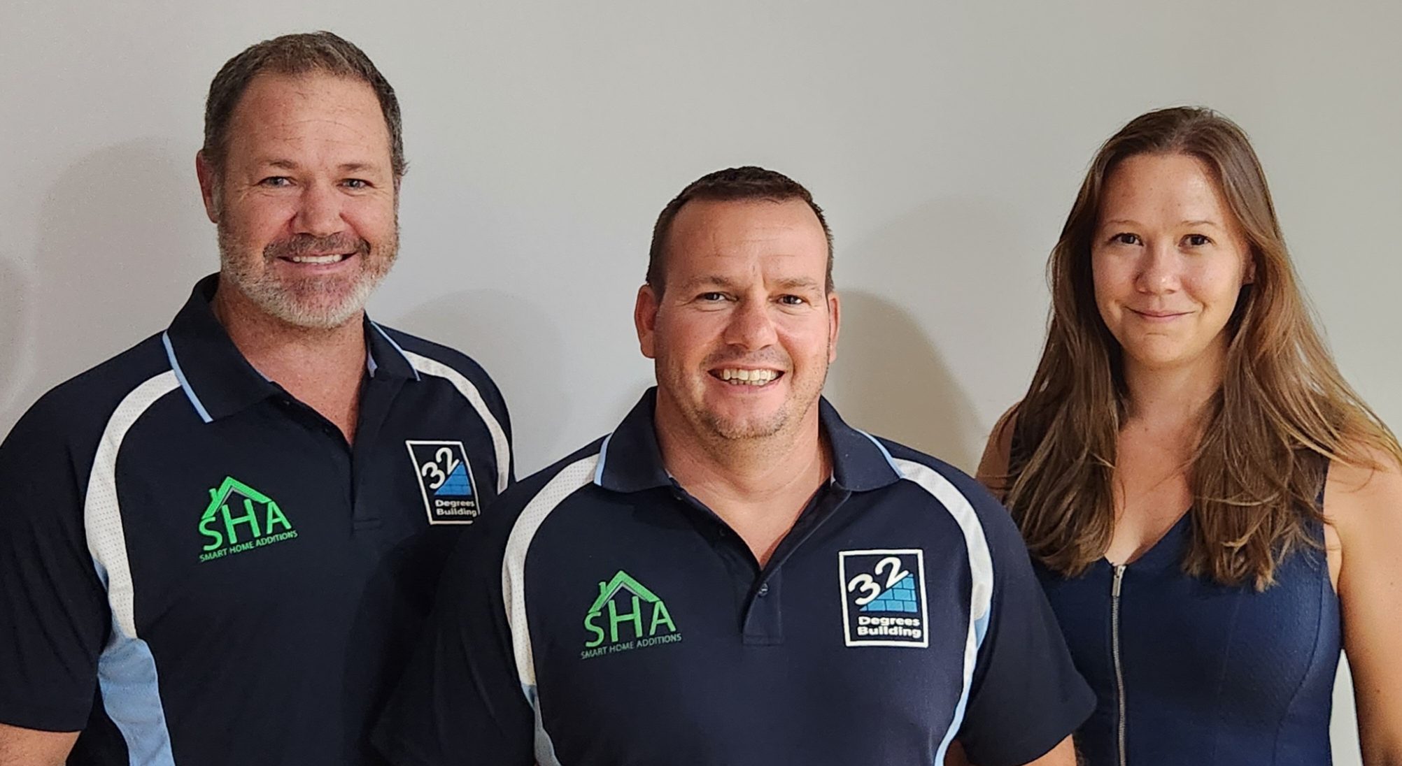 Denny, Alistair, Victoria Three people stand side by side smiling at the camera. Two men on the left wear matching dark shirts with company logos, while a woman on the right wears a sleeveless navy dress. The background is plain and light-colored.