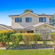 Second Storey Addition Narellan Vale (1 of 42) Two-story suburban house with beige siding, black-trimmed windows, and a dark roof, surrounded by trimmed hedges and green lawn under a bright blue sky. 32 Degrees Building logo in lower right corner.