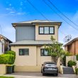 Second Storey Addition - Earlwood (2 of 50) A modern two-story beige house with a gray roof, driveway, and small front yard. A black car is parked in front, with neighboring houses on both sides under a blue sky.