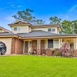Second Storey Addition - Narellan (4 of 34) A two-story brick house with a tiled roof, manicured lawn, shrubs, and a driveway with a black car. The house has large windows, a front porch, and is surrounded by trees under a blue sky.