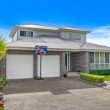 Second Storey Addition - Albion Park (1 of 33) A modern two-story brick house with a double garage, manicured lawn, small garden, and a basketball hoop on the driveway. The sky is clear and blue, and the property is surrounded by a metal fence.