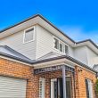 Second Storey Addition - Strathfield (2 of 32) Two-story modern house with a combination of red brick and white siding exterior, metal roof, large windows, and a garage. A blue sky and greenery are visible in the background.
