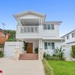 SECOND~3 A modern two-story white house with a balcony, green lawn, and plants by the entrance. The home has a paved driveway, stone pillars, and is situated between two neighboring buildings.